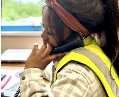 Woman wearing a high-visibility vest and headband, speaking on the phone at an office desk near a window.