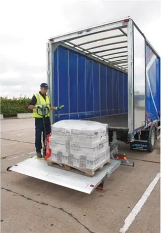 Logistics worker standing on the tail lift of a lorry, operating a pallet truck to unload a shrink-wrapped pallet of goods from an open-sided trailer.