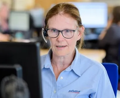 Customer service representative wearing a headset and glasses, working at a computer in a busy logistics office.