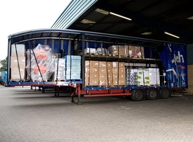 Double-deck curtain-side trailer fully loaded with pallets and various goods, shown at a distribution loading bay with the side curtain open for visibility.